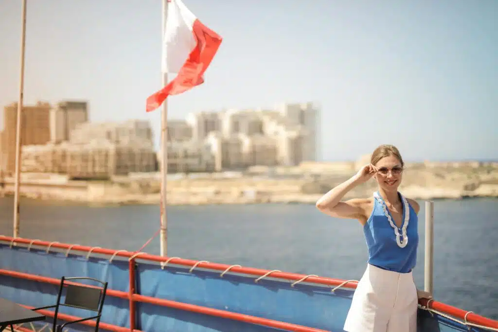 Smiling woman wearing sunglasses on a boat with a cityscape backdrop, enjoying a sunny day.