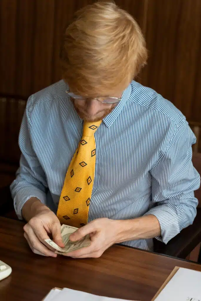 Red-haired businessman counting money at a desk, wearing a yellow patterned tie in an office setting.